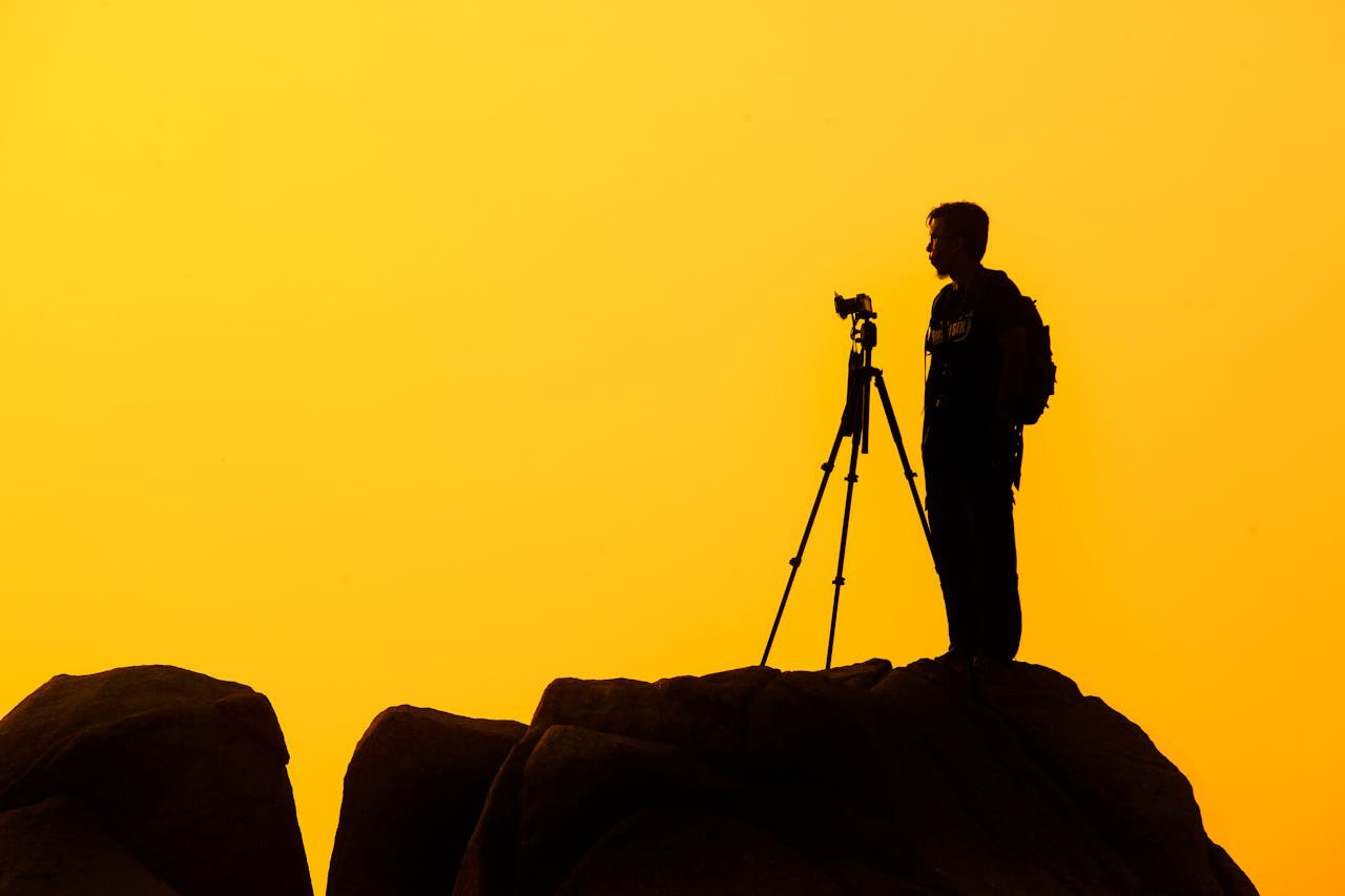 about-img-01 Silhouette of a lone photographer with tripod on a rocky cliff against a golden sunset sky.